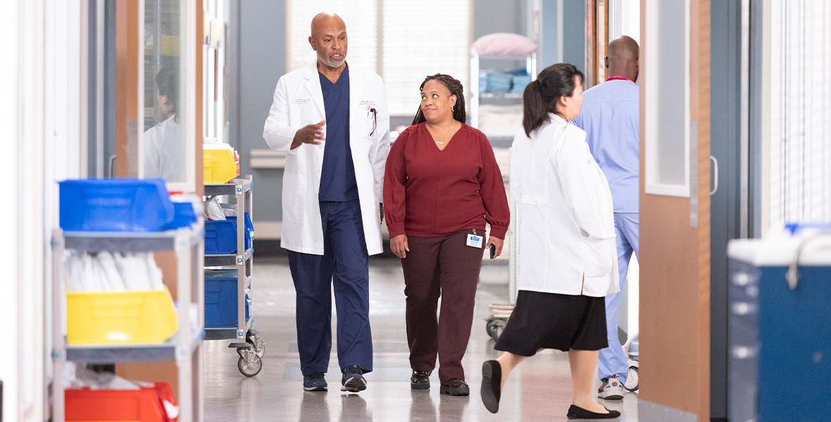 Actors James Pickens Jr. and Chandra Wilson walk down a long hallway in a hospital. Pickens Jr. wears a white lab coat over dark blue scrubs. Wilson wears a cardinal, long sleeved blouse and brown pants. A woman wearing a white lab coat and black skirt is seen walking into a room behind them. Metal carts with blue and yellow plastic containers line the hallway.