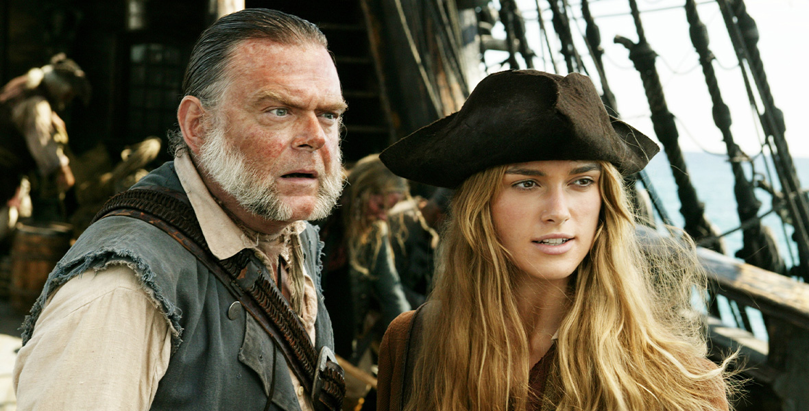 From left to right, actors Kevin McNally and Keira Knightley stand shoulder to shoulder. McNally wears a tan button-down shirt with a gray vest on top. Knightley wears a black tricorne hat and dark brown jacket. Behind them are large ropes and the ship’s cabin.