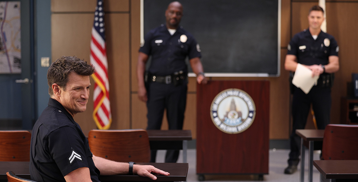 Actor Nathan Fillion smirks and sits in a wooden chair next to a long wooden desk. He wears a black cop uniform with two white chevrons embroidered on his right sleeve. His left arm rests on the top of the desk and his right arm rests on his right thigh. At the front of the room is an American flag along with a large wooden podium and a large chalkboard hanging on the wall. Two male officers stand on each side of the podium and look at Fillion.