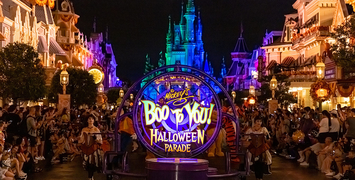A round purple sign is held up during the parade. In yellow letters, the sign reads “Boo-To-You! Halloween Parade.” Main Street, U.S.A., is lit up in warm white lights as guests gather around the parade.