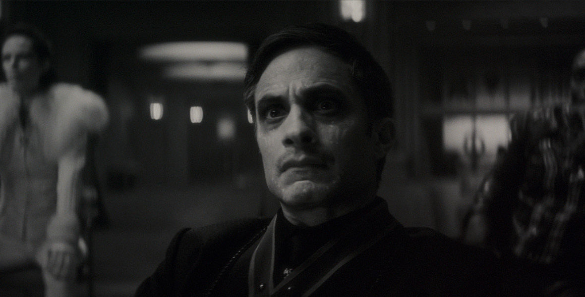 In a still from Werewolf by Night, actor Gael García Bernal portrays Jack Russell; he wears all-black clothing and sits in a chair. Behind him is a person dressed in white with a fur stole around their shoulders.