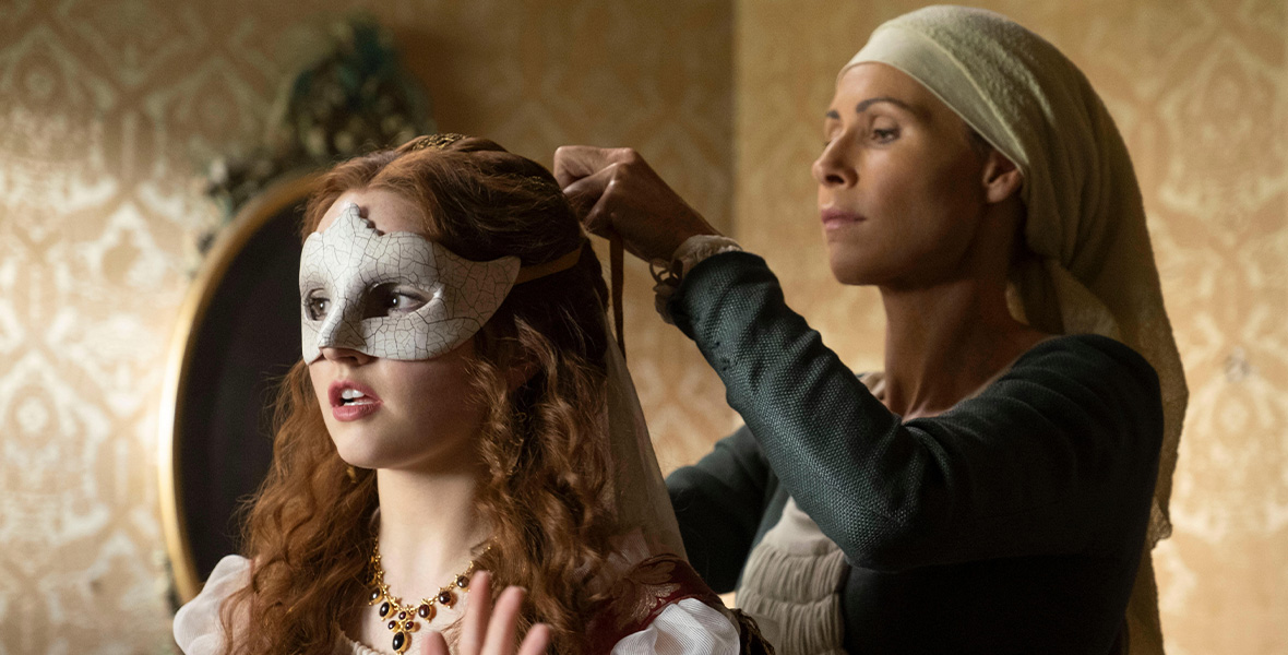 From left to right, Kaitlyn Dever stands with her hands raised. She wears a white mask covering the top half of her face. She wears a red velvet dress with white puffy sleeves and gold embroidered accents. Actor Minnie Driver wears a white head scarf with a long-sleeved blue dress. She stands behind Dever and ties the mask onto her head.
