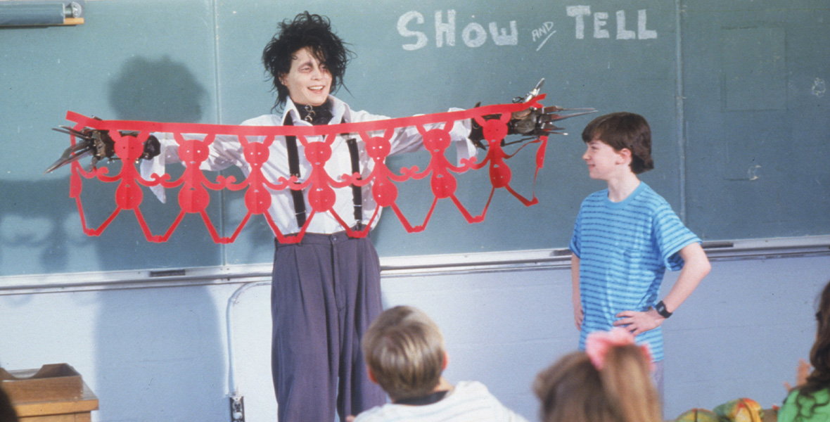 Actor and Disney Legend Johnny Depp portrays Edward Scissorhands. He stands with his arms extended outward and holds a long strip of red paper cut into a design. Depp wears a black turtleneck under a white, button-down dress shirt. He wears dark gray pants that are held up by black suspenders. His hands are comprised of metal blades. A young boy stands to his left with his hands placed on his hips. He smiles and looks on at Depp. He wears a blue shirt with dark blue stripes. Behind them is a green chalkboard reading, “Show and Tell.” In front of them, young children sit in desks facing them.