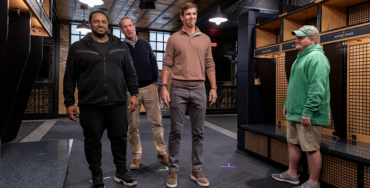 From left to right, former American football players Jerome Bettis, Peyton Manning, and Eli Manning stand inside the football team’s locker room at the University of Notre Dame. On the right stands a man who wears a green sweatshirt with a matching hat and tan cargo shorts. Bettis wears a black tracksuit with a black T-shirt. Eli wears a light brown quarter-zip sweater with the sleeves pushed up, and blue denim jeans. Peyton pumps his right fist. He wears a navy-blue quarter-zip sweater with tan pants.