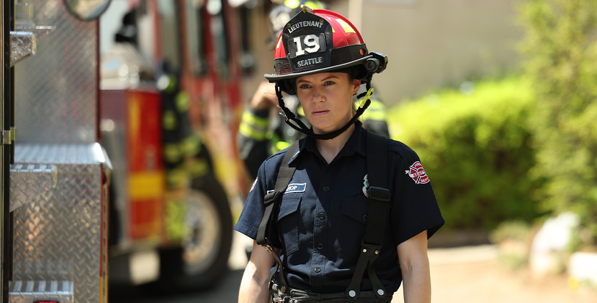 Actor Danielle Savre stands adjacent to a large red firetruck. She wears a red firefighter hat that reads “Lieutenant 19 Seattle” with navy blue uniform shirt and black slacks with black suspenders. Additional firefighters are seen in the background.