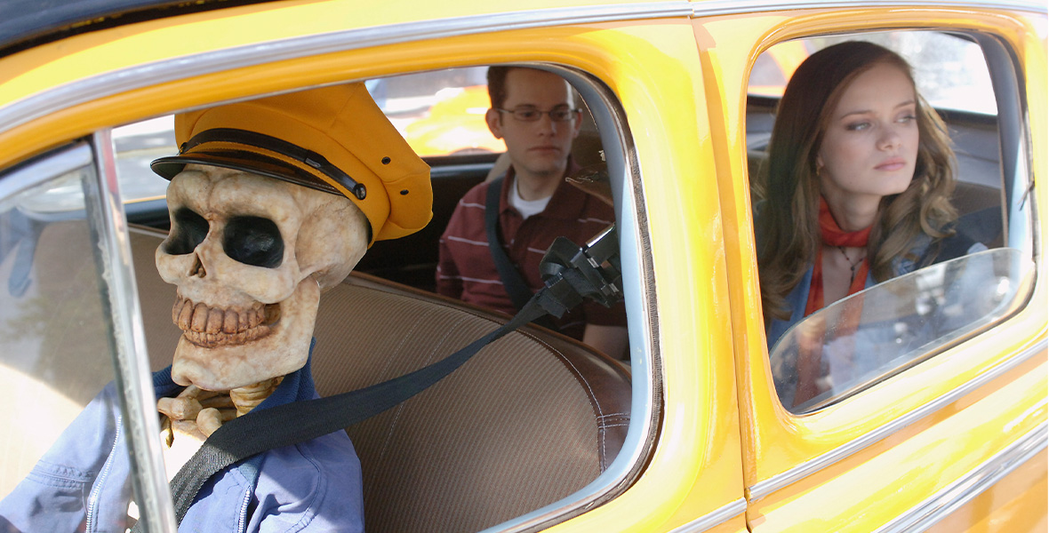 A skeleton sits in the driver’s seat of a vintage yellow cab. It wears a yellow hat and dark blue Member’s Only jacket. Seated in the backseat are actors Joey Zimmerman and Sara Paxton. Zimmerman wears a white-striped cardinal polo shirt over a white T-shirt. Paxton wears a blue denim jean jacket with an orange scarf wrapped around her neck. Zimmerman and Paxton both look to the left with the window rolled down.