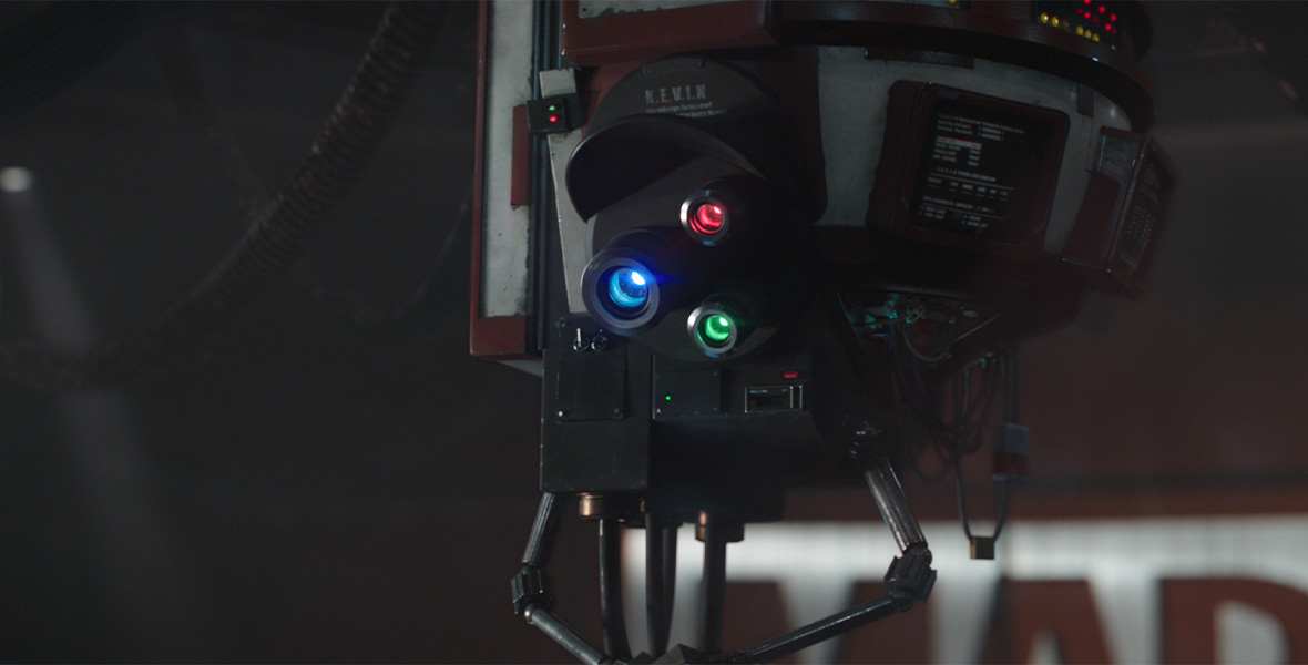 A robotic machine suspended from the ceiling with three lenses colored blue, red, and green. The machine reads, “K.E.V.I.N.” Behind the machine, part of the Marvel Studios logo is seen in red and metallic silver.