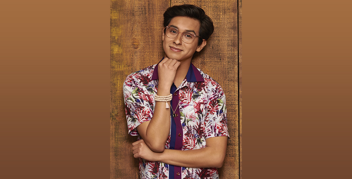 Frankie A. Rodriguez stands against a wood-paneled wall with his right hand resting underneath his chin. He is wearing a purple, white, and blue floral patterned top and clear-frame eyeglasses.