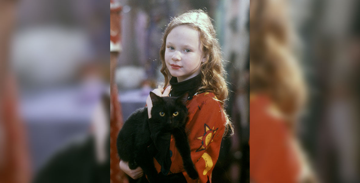 Dani from the film Hocus Pocus holds Binx, a black cat, in her arms. They are both facing the camera and looking at the viewer.