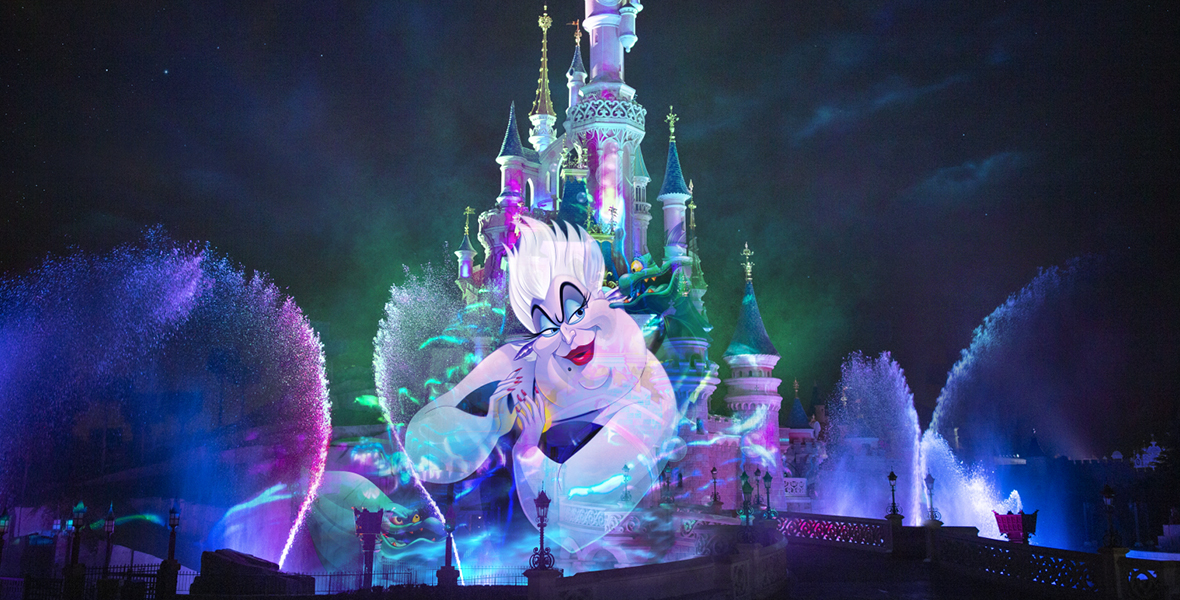 Ursula the sea witch from The Little Mermaid is projected onto the Disneyland Paris castle. Large gusts of water are splashing around the imagery. Ursula has short white hair, blue eyeshadow, and red lipstick.