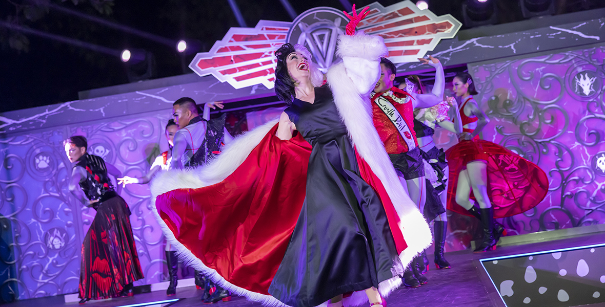 Cruella de Vil wears a black satin dress with a large red lined, white fur jacket and red gloves. She is standing at an angle with her left hand in the air.