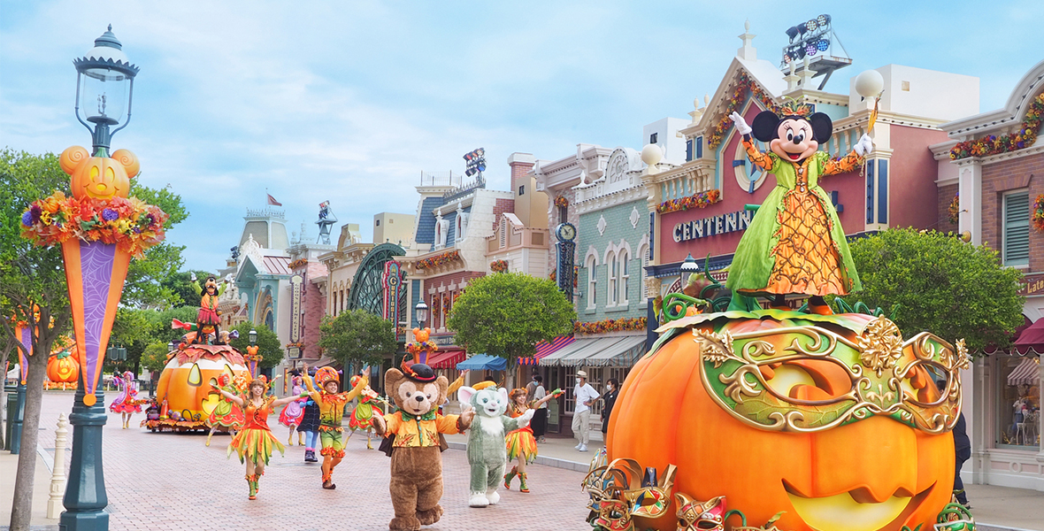 Minnie Mouse stand atop an orange pumpkin float leading a parade down Main Street, U.S.A. She wears an orange dress with green overlay. She is followed by several parade performers in matching color schemes.