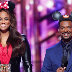 Tyra Banks wears Minnie Mouse ears, which include a red bow with white polka dots; a black leather jacket with a studded lapel; an embellished, oversized belt buckle; and a red satin dress. She is smiling and holding a microphone in her right hand. To her left is Alfonso Ribeiro, who wears Mickey Mouse ears and an all-navy suit.
