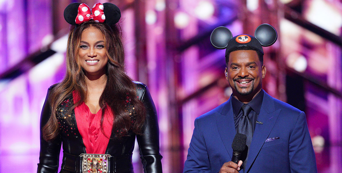 Tyra Banks wears Minnie Mouse ears, which include a red bow with white polka dots; a black leather jacket with a studded lapel; an embellished, oversized belt buckle; and a red satin dress. She is smiling and holding a microphone in her right hand. To her left is Alfonso Ribeiro, who wears Mickey Mouse ears and an all-navy suit.