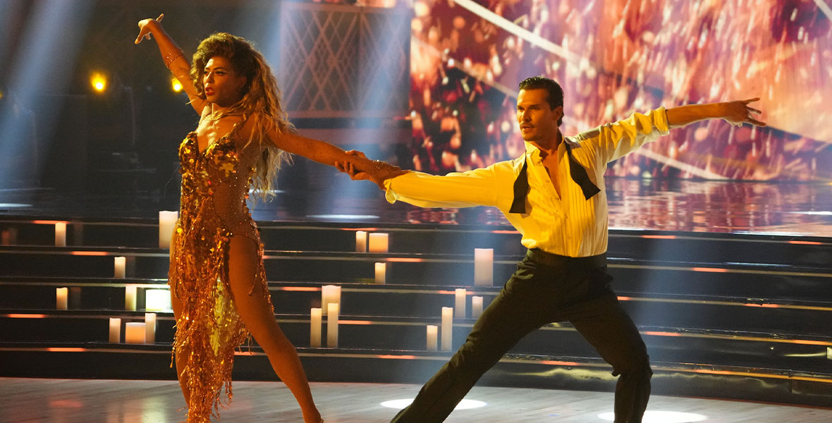 Shangela wears a gold fringe dress with a thigh-high slit. Her right arm is extended in the air, and she is holding onto Gleb Savchenko, who is to her left, with her left arm. Savchenko’s legs are spread wide, and his left arm is extended. He is wearing a partially unbuttoned white dress shirt, black pants, and an undone bow tie.