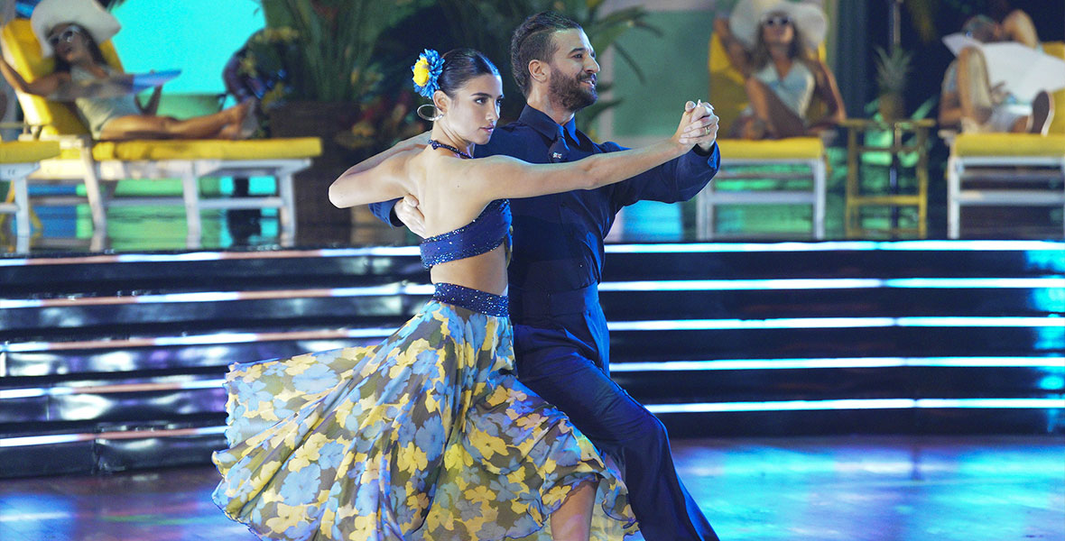 Charli D’Amelio and Mark Ballas hold a classic tango pose as they glide across the dance floor. D’Amelio is wearing a midriff-baring, bedazzled black top, a flowing floral skirt, and black and white dance shoes. She has a yellow and blue flower in her hair. Ballas is wearing a black dress shirt, black pants, and black and white dance shoes.