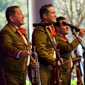 Three Mariachi singers are in front of three black stand microphones at EPCOT. The singer on the left is holding a trumpet and has short dark hair; the one in the middle is holding a brown violin and has short dark hair; and the on the right is holding the black microphone, a brown violin, and has short dark hair. All are smiling wide. Each is wearing a forest green suit embroidered with gold and yellow detailing, with white shirts underneath and have a red and gold bowtie. Behind the main three are additional Mariachi band members.
