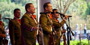 Three Mariachi singers are in front of three black stand microphones at EPCOT. The singer on the left is holding a trumpet and has short dark hair; the one in the middle is holding a brown violin and has short dark hair; and the on the right is holding the black microphone, a brown violin, and has short dark hair. All are smiling wide. Each is wearing a forest green suit embroidered with gold and yellow detailing, with white shirts underneath and have a red and gold bowtie. Behind the main three are additional Mariachi band members.