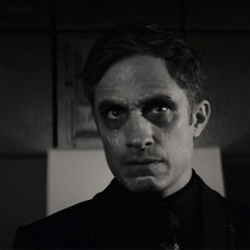 Gael García Bernal, in character as Jack Russell, has white markings on his face. He is standing in a dimly lit room and has a stern look on his face.