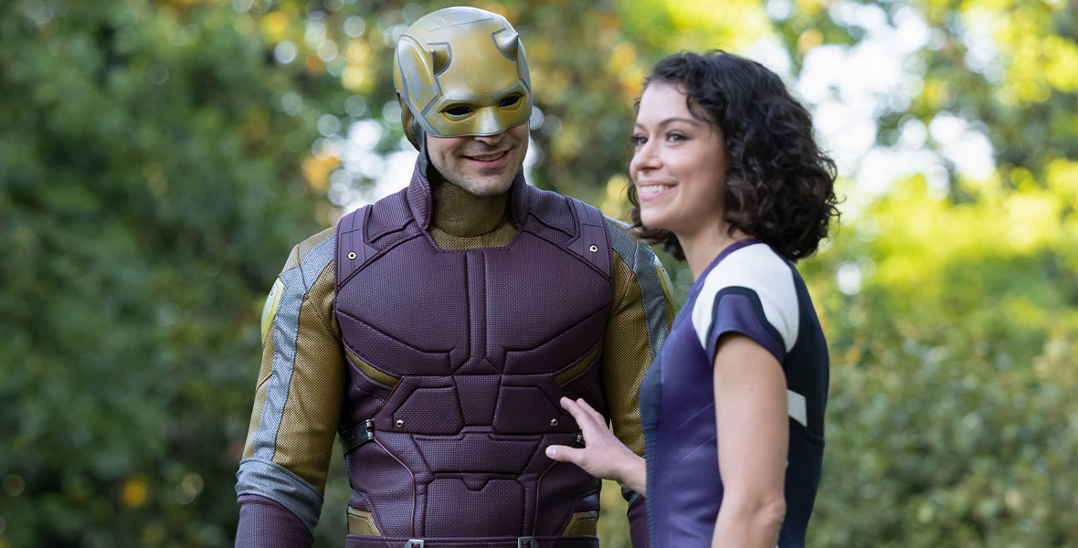 From left to right, Daredevil wears a maroon and gold costume and smiles. Jennifer Walters, wearing a purple and white super suit, has shoulder-length curly brown hair. She is smiling and lifting her right arm towards him. Behind them are trees.