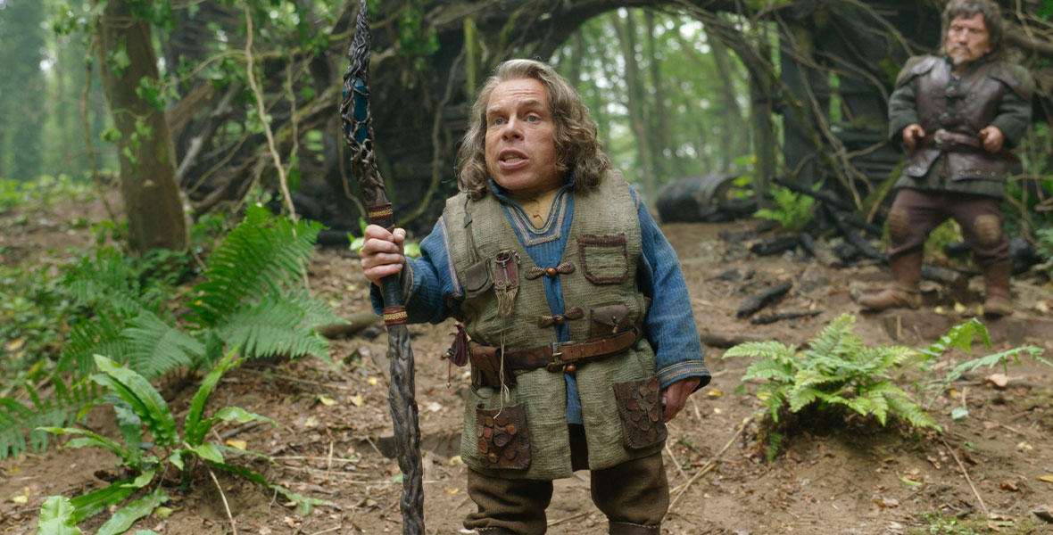 Warwick Davis, in character as Willow Ufgood, holds a staff as he stands in a forest. He has shoulder-length hair and is wearing a green vest with multiple pockets.