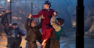 In the center, Mary Poppins, played by Emily Blunt, wears a red jacket and a navy blue hat with matching shoes, in a still from Mary Poppins Returns. She is being lifted into the air by three singing lamplighters, while other lamplighters dance around them at nighttime in the street.