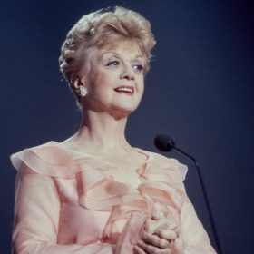 Angela Lansbury stands onstage in front of a blue curtain, wearing a frilly pink gown.