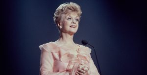 Angela Lansbury stands onstage in front of a blue curtain, wearing a frilly pink gown.