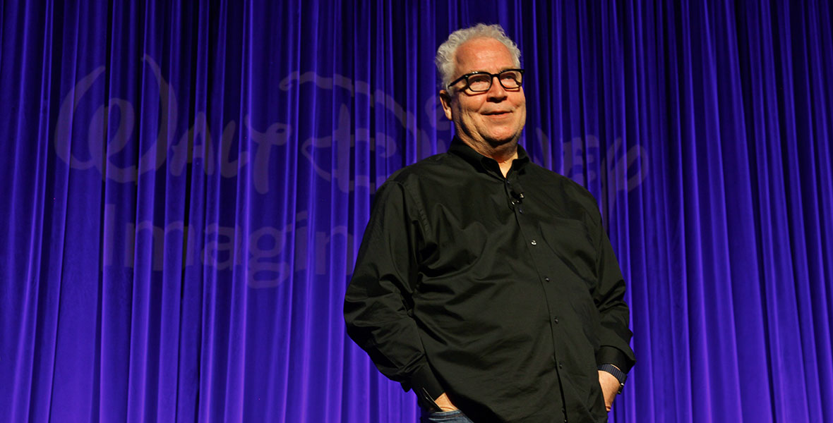 Bob Weis, Global Imagineering Ambassador and former President of Walt Disney Imagineering, stands alone on a stage in front of a blue curtain. The logo for Walt Disney Imagineering is projected on the curtain behind him.