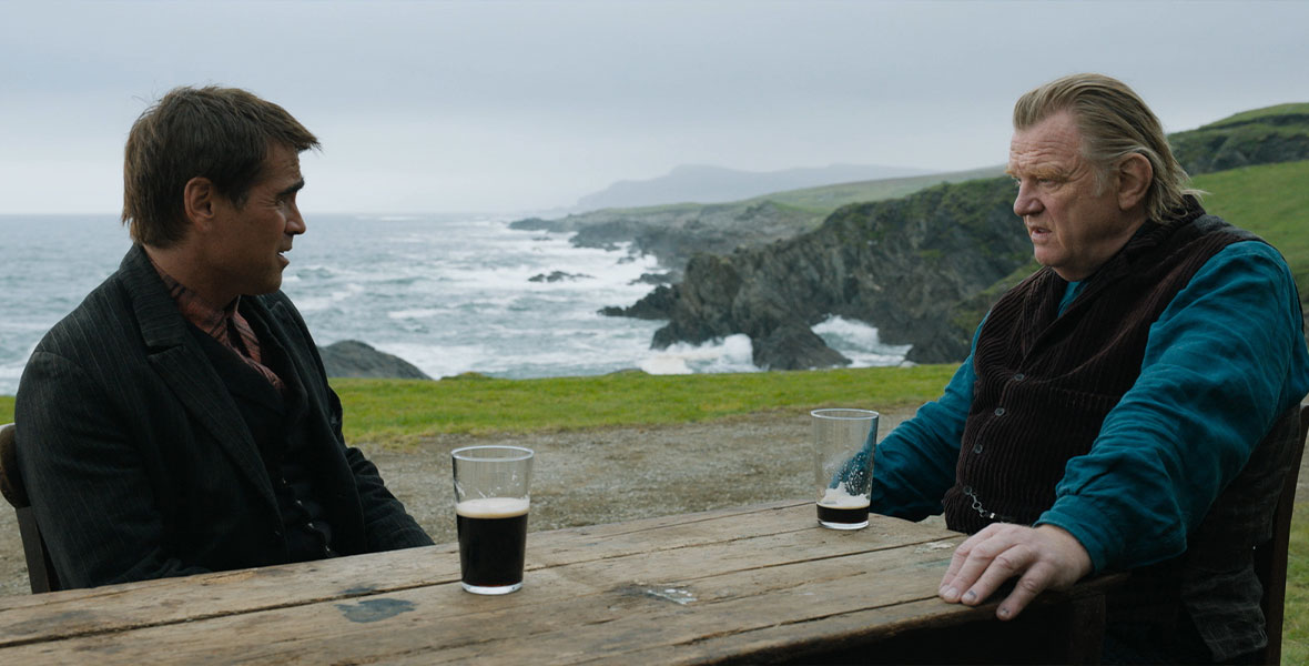 Colin Farrell (left) and Brendan Gleeson (right) are sitting outside on the Irish coastline and sharing a pint. They are facing each other and having a conversation. Behind them are rolling green hills. Waves crash into rocks. The weather is overcast.