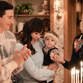 From left to right actors Sam Lerner, Hayley Orrantia, and Wendi McLendon-Covey stand around a wooden table. Lerner wears a tan sweater with black abstract shapes and claps his hands. Orrantia wears a light blue sweater with black pants. She holds an infant girl in her arms. The infant wears a black jumper and animal print onesie. McLendon-Covey wears a black sweater with large colorful-sequined dreidels and a white blouse. On the table is a gold menorah lit next to a glass of red wine and a small blue candle.