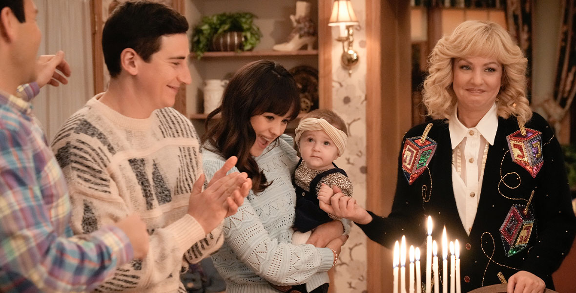 From left to right actors Sam Lerner, Hayley Orrantia, and Wendi McLendon-Covey stand around a wooden table. Lerner wears a tan sweater with black abstract shapes and claps his hands. Orrantia wears a light blue sweater with black pants. She holds an infant girl in her arms. The infant wears a black jumper and animal print onesie. McLendon-Covey wears a black sweater with large colorful-sequined dreidels and a white blouse. On the table is a gold menorah lit next to a glass of red wine and a small blue candle.