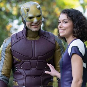 Charlie Cox as Daredevil and Tatiana Maslany as Jennifer Walters in a still from She-Hulk: Attorney at Law. Daredevil is in his full superhero outfit which looks like tactical gear in a deep purple-red, with gold accents. He is wearing his iconic gold devil helmet face mask. Jennifer is in her regular human form and wearing her purple and white She-Hulk jumper; she is looking off camera and Daredevil is looking at her, smiling.