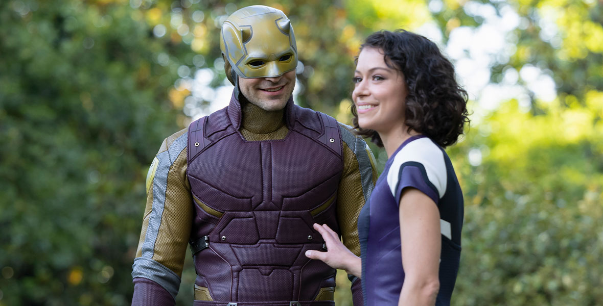 Charlie Cox as Daredevil and Tatiana Maslany as Jennifer Walters in a still from She-Hulk: Attorney at Law. Daredevil is in his full superhero outfit which looks like tactical gear in a deep purple-red, with gold accents. He is wearing his iconic gold devil helmet face mask. Jennifer is in her regular human form and wearing her purple and white She-Hulk jumper; she is looking off camera and Daredevil is looking at her, smiling.