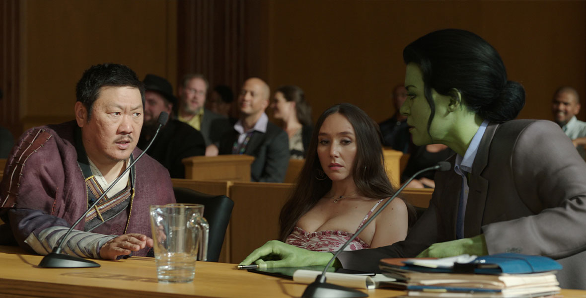 Wong sits in court with Madisynn to his right and Jen Walters to her right. He is wearing his Kamer Taj robes and looking concerned. Madisynn is staring off into space and she is wearing a tight pink floral dress. Jen is in her She-Hulk form, wearing a suit, and looking back at Wong as if discussing her next move.