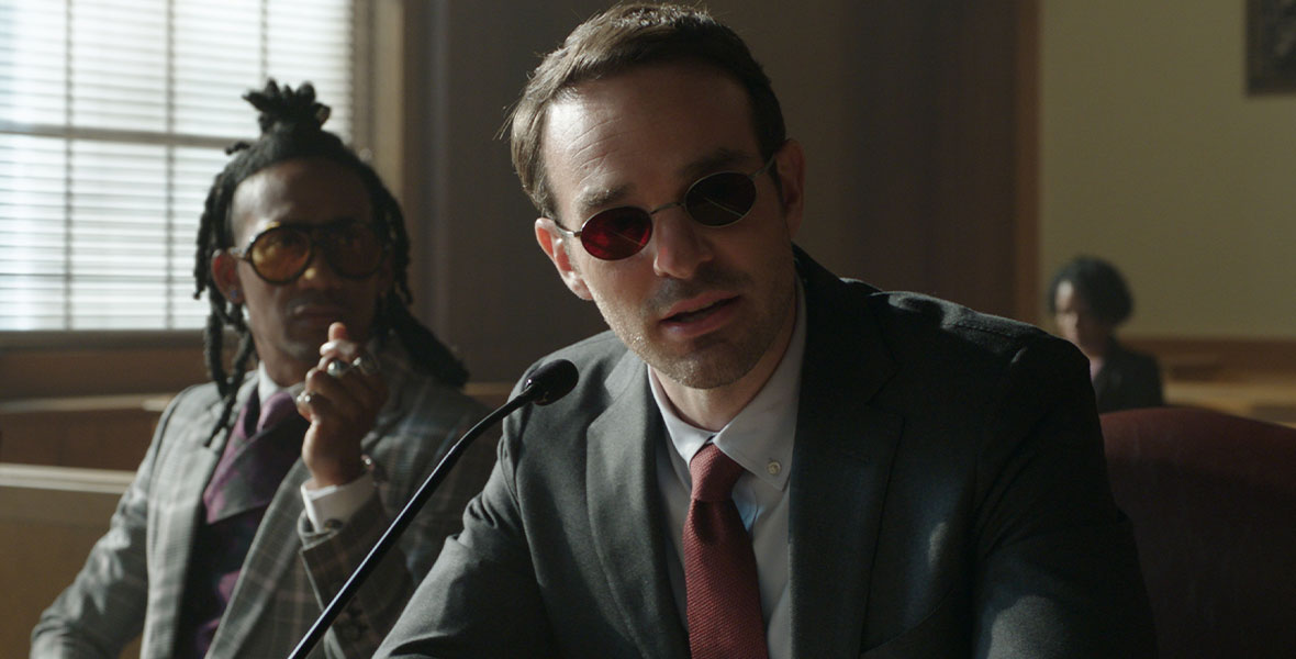 Matt Murdock in his normal clothes (a gray suit with a red tie) sits in court with his client, Luke Jacobson. Jacobson’s hair is tied up in a knot on his head and he is wearing a plaid suit and orange sunglasses. Murdock is speaking into a microphone in front of him.