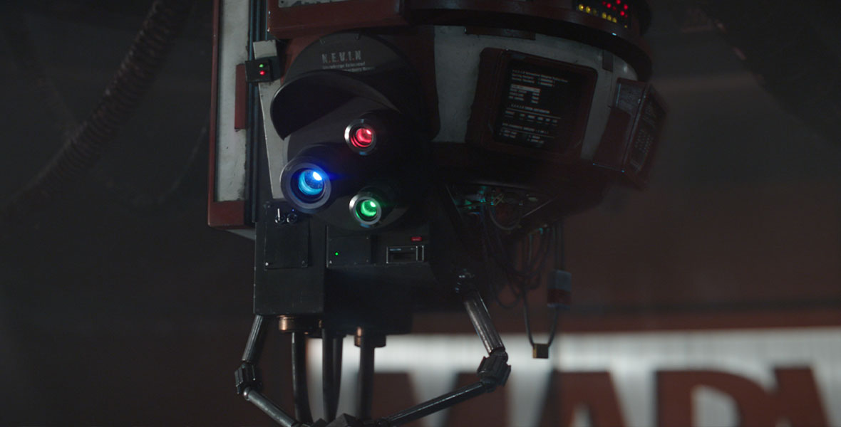 A still of the robot, K.E.V.I.N. This bot is suspended from the ceiling and has three lights on its “face”, one blue, one red, and one green. There is a bill above the lights that resembles a baseball cap. Above the “cap” is white writing that reads “K.E.V.I.N.”