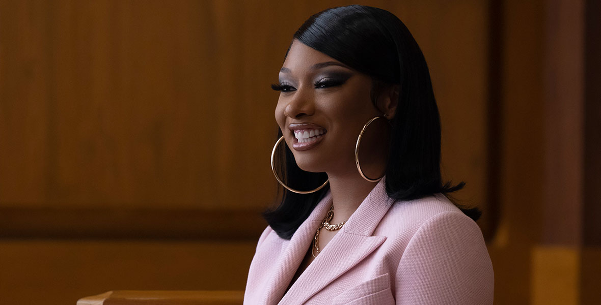 Megan Thee Stallion, as herself, in She-Hulk. Megan is wearing a pink blazer, gold chain necklaces, and large gold hoop earrings. Her hair is parted on the side and slick straight. She is in the courtroom, but looking very happy with a big smile on her face.
