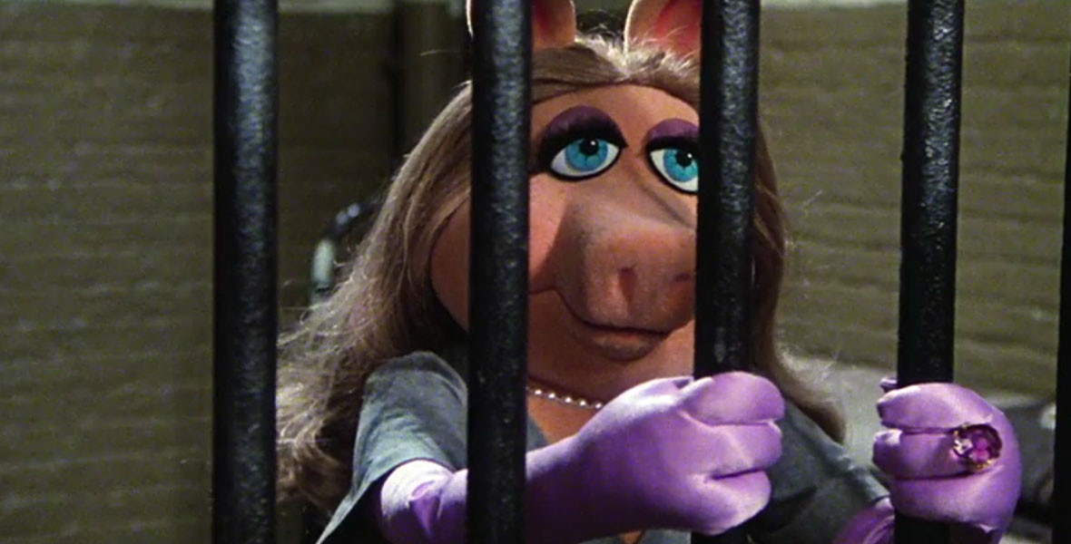 Miss Piggy in The Great Muppet Caper. She is behind bars in a jail cell and wearing long, purple satin gloves, a blue dress, and a pearl necklace.