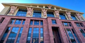 The Team Disney Building at The Walt Disney Studios lot in Burbank, California is red brick with a tan roof. Toward the top of the building are huge statues of seven dwarfs from Snow White, holding up the roof. Six are spread out equidistant to one another between the third and fourth story, Dopey is centered on the floor above the rest with his hands up as if holding up the peaked roof. The sun is shining behind the building with a bright blue sky.
