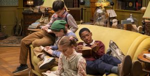 From left to right, The Mysterious Benedict Society actors Mystic Inscho, Emmy DeOliveira, Seth Carr, and Marta Kessler sit on a yellow couch, each immersed in a book.