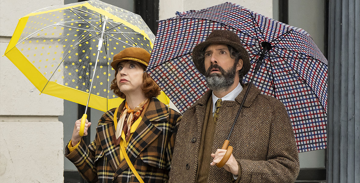 In a still from The Mysterious Benedict Society, from left to right, actors Kristen Schaal and Tony Hale hold patterned umbrellas and wear thick wool coats. They are both looking up and appear shocked.