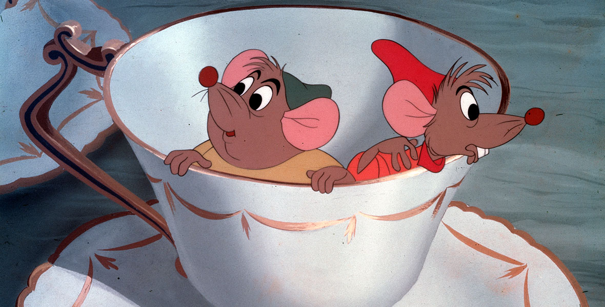 In a still from Cinderella, Gus, a mouse in a green hat and yellow shirt, and Jaq, a mouse in a red hat and orange shirt, peer out of a white and gold teacup.