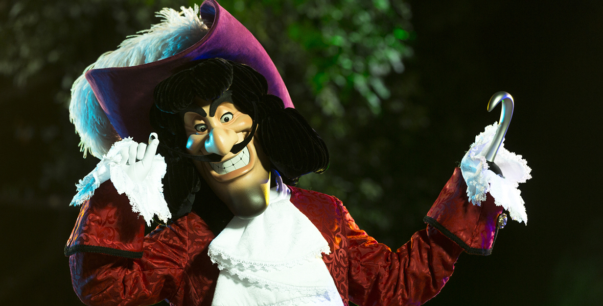 Captain Hook stands with his right hand and left hook in the air. His head is tilted to the right. He is wearing a red jacket with a frilly white shirt underneath. He has long, wavy black hair and a mustache. He is wearing a purple captain’s hat with a large white feather.
