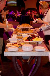 Members going through the breakfast line filling up their plates. The breakfast table is white with silver legs. On the table are platters of fruit, eggs, and pastries. In the background, multiple guests are mixing and mingling in the space.