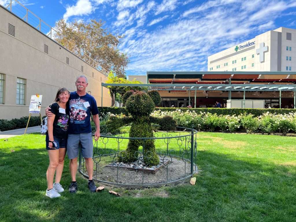 Two guests posing next to the Mickey horticulture bush. The guest on the left has long brown hair and is wearing a black T-shirt with neon designs and blue jean shorts. This guest is hugging the guest to the right. The guest on the right has short gray hair, a mustache and is wearing a navy-blue shirt with a red train on it and grey shorts. To the right of the guests is a bush trimmed to look like Mickey Mouse. The guests are standing on a green lawn.