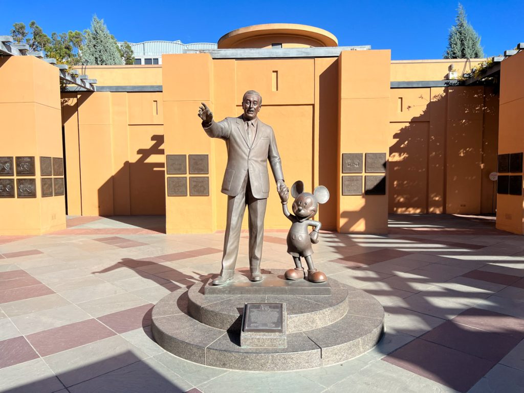 Partners statue surrounded by a tan building and grey and red flooring. The Partners statue is in bronze and is of Walt Disney walking with and holding Mickey Mouse’s hand.