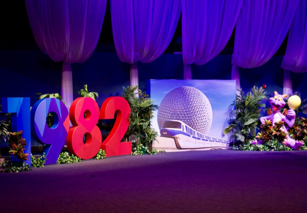 At a recent EPCOT 40th event, on the left side of a photo op are large numbers reading “1982.” The 19 is in blue and the 82 is in red; surrounding the numbers are green leaves, trees, and bushes. To the right of the numbers is another photo background of Spaceship Earth during the day, with the white and blue monorail driving past. To the right of that image is a large purple Figment statue holding a yellow balloon, with green leaves, trees, and bushes. Above the photo opportunities is white fabric draped from the ceiling. The room is illuminated with blue and purple lights.