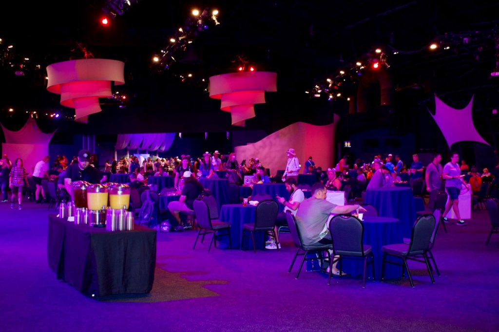 Large banquet room full of round tables, with black tablecloths and black chairs, at a recent EPCOT 40th event. People are sitting at tables and walking around. The room is highlighted with pink and purple lights. Hanging from the ceiling are spiraled white lamps. The left side of the photo features the drink table with pitchers or orange juice, apple juice, and water.