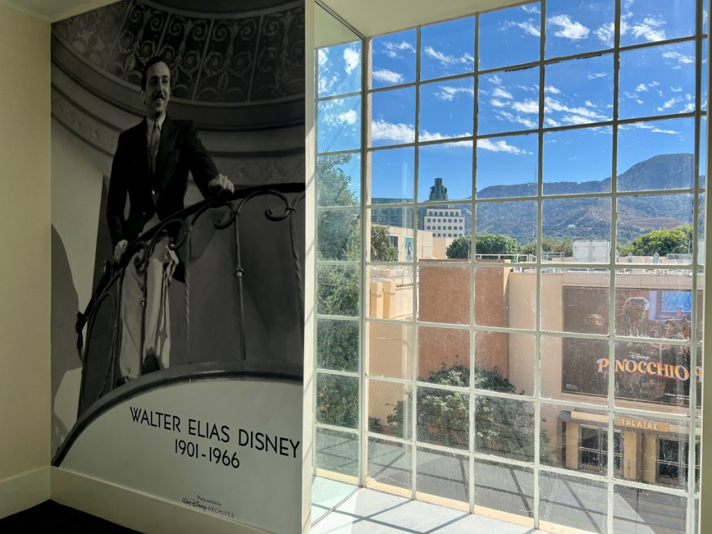 Long large black and white photo of Walt Disney standing on a balcony. Walt is wearing a suit and tie. On the photo in the bottom left corner states Walter Elias Disney and below that, 1901-1966. To the right of the photo is a window looking out to a blue sky and the Studio Theatre.